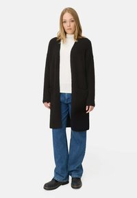 Schwarzer langer Cardigan mit offenem Front, Taschendetails und gerippter Textur, kombiniert mit einem weißen Rollkragenpullover und blauen weiten Jeans.