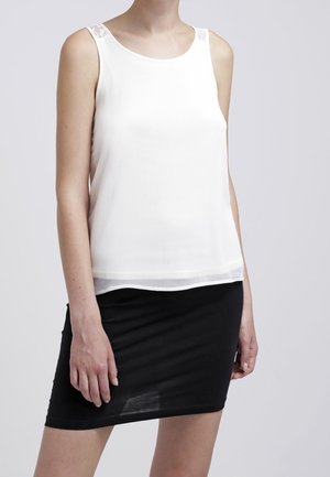 Mujer vistiendo una blusa blanca sin mangas con detalles de encaje y una falda negra ajustada frente a un fondo liso.