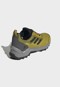 Vihreä ja harmaa juoksukenkä, jossa on teksturoitu verkkomateriaali, mustia yksityiskohtia, paksu kumipohja ja kirkkaat oranssit nauhat.