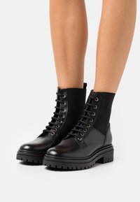 Botas negras de tobillo hechas de cuero con cordones en la parte delantera, paneles laterales elásticos y suelas de goma gruesa. Presentan ojales metálicos y cuero texturizado.
