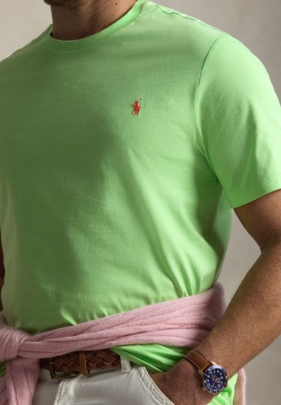 Ανοιχτό πράσινο κοντομάνικο μπλουζάκι με ένα μικρό κόκκινο κεντημένο λογότυπο, σε συνδυασμό με λευκό παντελόνι και ένα ροζ πουλόβερ δεμένο γύρω από τη μέση.