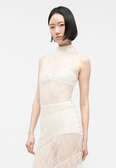 Νεαρή γυναίκα με κοντά μαύρα μαλλιά που φοράει λευκό διάφανο δαντελωτό αμάνικο φόρεμα με ψηλό γιακά και κουμπιά στη μία πλευρά.