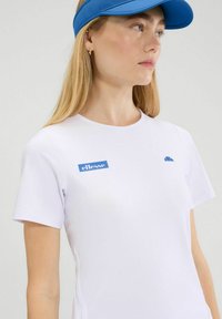 Camiseta deportiva de manga corta blanca con el logo "ellesse" en azul y un acento de nubes. Textura suave, cuello redondo, diseño minimalista.