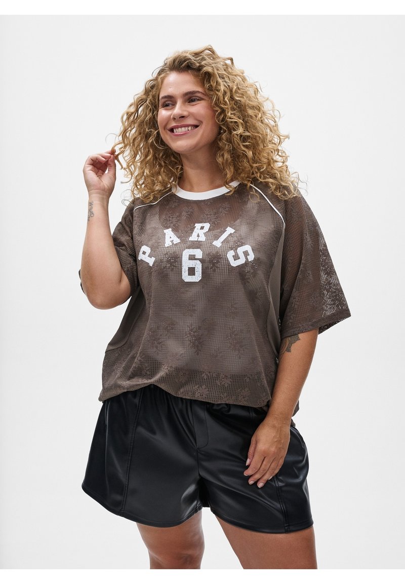 Mujer sonriente con cabello rizado y rubio que lleva un top marrón de malla con "Paris 6" y pantalones cortos de cuero negro, posando contra un fondo blanco.
