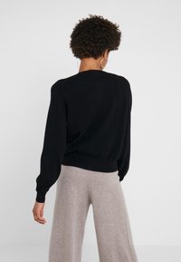 Czarny sweter z miękkiej, teksturowanej tkaniny, z balonowymi rękawami i prążkowanym dołem. Połączony z szarymi, szerokimi spodniami. Widok z tyłu.