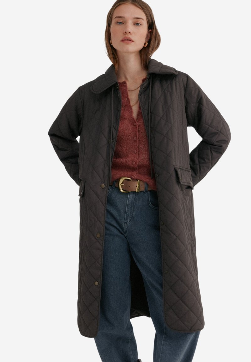 Gequilted zwarte buitenkleding met een kraag, drukknoopsluiting, twee voorzakken en een middellange pasvorm. Gedeeltelijk over een rode gebreide trui gedragen.