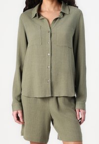 Chemise boutonnée vert olive à manches longues, avec deux poches poitrine, texture douce et léger éclat, assortie à un short coordonné.