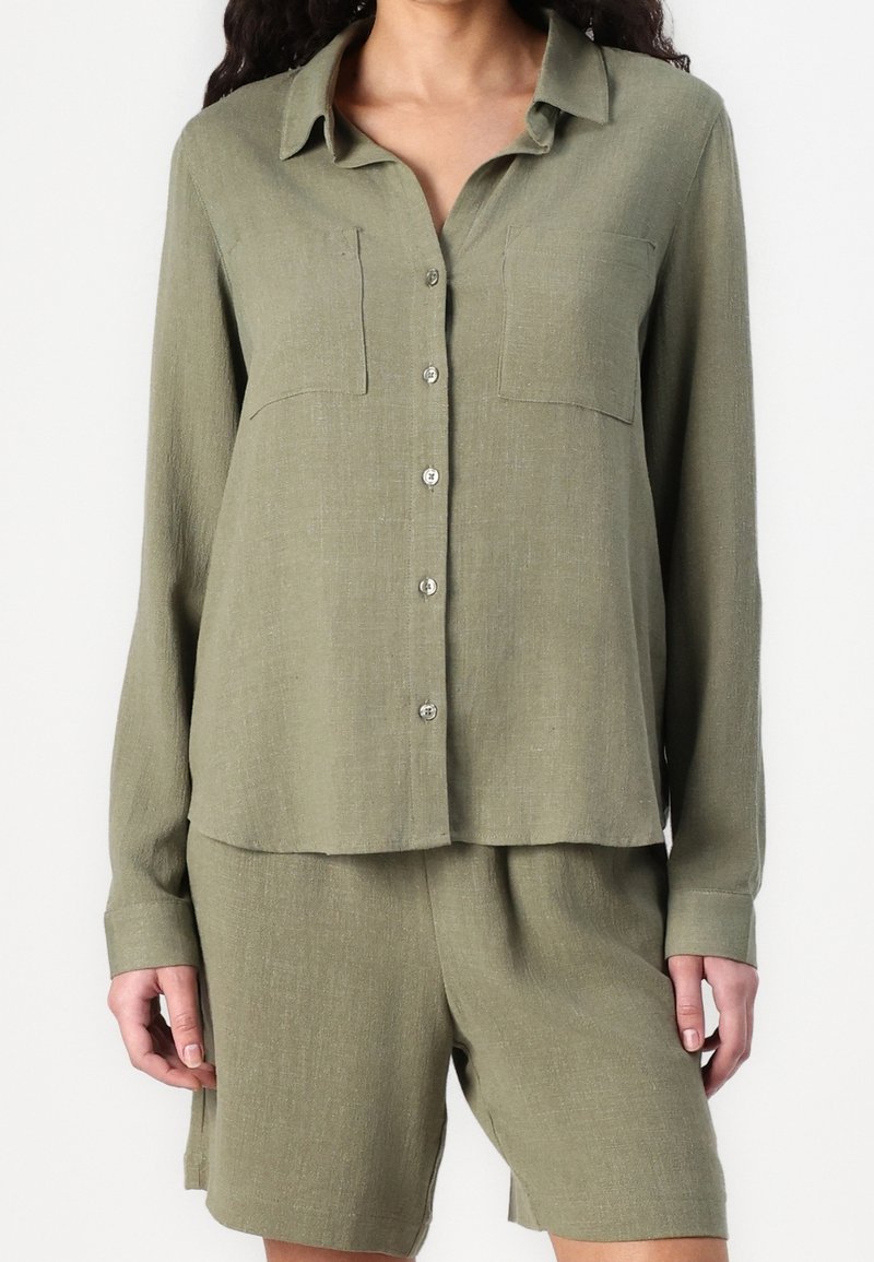 Chemise boutonnée vert olive à manches longues, avec deux poches poitrine, texture douce et léger éclat, assortie à un short coordonné.