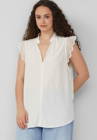 Blouse blanche sans manches avec un décolleté en V et des détails en dentelle sur les épaules. Tissu doux avec une coupe décontractée et un ourlet arrondi.