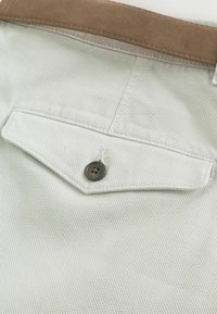 Gros plan sur la poche d’un pantalon en tissu texturé beige, dotée d'un rabat boutonné, avec une passoire de ceinture en daim marron visible au-dessus.