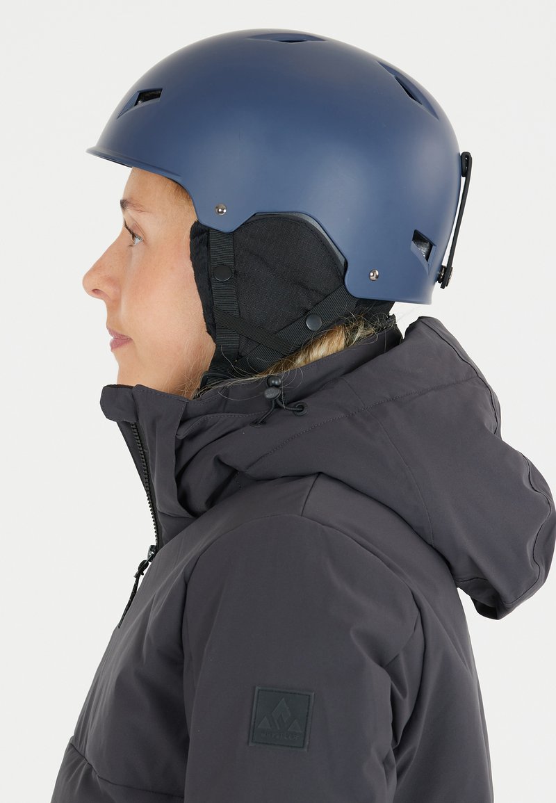 Marinstrids hjälm för skidåkning med ventilationshål, justerbara remmar och öronkuddar av svart tyg, som bärs med en mörkgrå jacka som har en logotyp-patch.