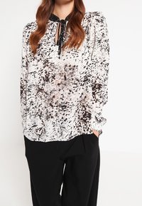 Blusa a maniche lunghe con motivo bianco e nero, colletto a occhiello e dettagli con cravatta nera, abbinata a pantaloni neri.