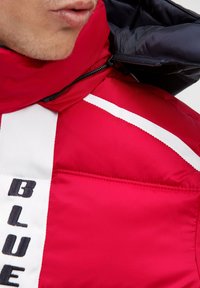 Red padded jacket with a high collar, featuring contrasting white stripes and a black zip. "BLUE" text in black on the front.