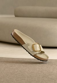 Μπεζ σανδάλι με πλατιά λουρί, μεγάλη χρυσή αγκράφα και υφασμένο φελλό στο πέλμα· μαύρη λαστιχένια σόλα με λεπτομέρειες αντιολισθητικού.
