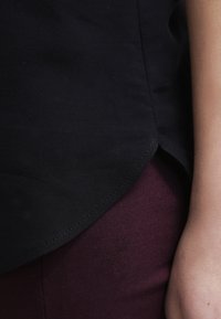 Musta kangaspaita, jossa on kaareva helma ja sileä pinta. Beige käsi lepää marooniväristen housujen vieressä, mikä näyttää vaatteiden istuvuutta.