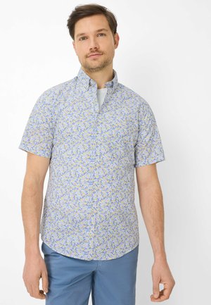 Man wearing a short-sleeve button-up shirt with a blue and yellow floral pattern and light blue pants, standing against a white background.