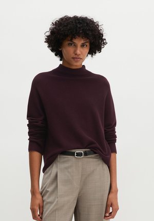 Vrouw met kort krullend haar draagt een donkerpaarse trui, ingestopt in beige geplooide broek met een zwarte riem tegen een effen witte achtergrond.