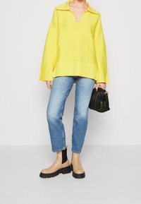 Personne portant un pull ample en V jaune, un jean bleu clair, des bottes chunky beiges, tenant un sac à main noir texturé devant un fond neutre.