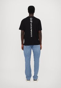Svart bomulls T-skjorte med vertikal hvit tekst som leser "EA7 EMPORIO ARMANI" på baksiden, kombinert med lys blå jeans og sneakers.