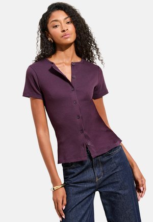 Frau mit lockigem Haar, die ein enganliegendes, kurzärmeliges lila Hemd mit Knöpfen und dunkelblaue Jeans trägt, posiert mit einer Hand in der Hüfte.