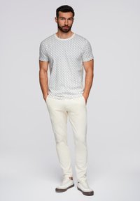 Ein bärtiger Mann, der ein weißes, gemustertes T-Shirt, hellbeige Hosen und weiße Sneakers trägt, steht mit den Händen in den Taschen vor einem einfarbigen hellen Hintergrund.