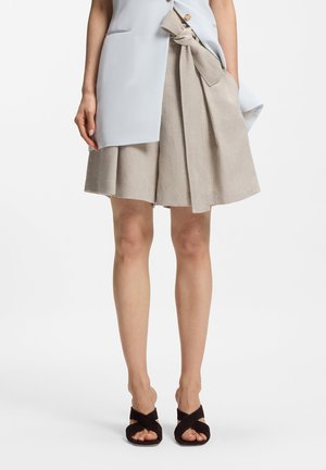 Kobieta ubrana w beżową plisowaną spódnicę z wiązaniem w talii, jasnoniebieską marynarkę oraz czarne sandały na obcasie z odkrytymi palcami, stoi na tle białego tła.