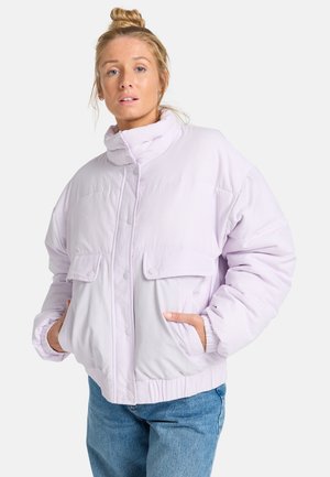 Kobieta o blond włosach związanych w kok, ubrana w jasnofioletową puchową kurtkę z rękami w bocznych kieszeniach i niebieskie dżinsy na jednolitym tle.