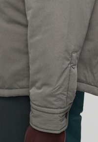 Γκρι, υφασμάτινο μανίκι σακακιού με ραμμένο μανσέτα και κρυφό κουμπί, συνδυασμένο με σκοτεινό μπλε ένδυμα στο φόντο.