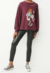 Sweatshirt bordeaux avec des manches balloons, comportant un graphique d'un personnage tenant des cadeaux et le texte "Joyeuses Fêtes". Associé avec des leggings noirs et des baskets grises.
