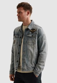 Muž v svetlomodrej rifľovej bunde s vyšívanými nášivkami, pod bundou béžová košeľa a čierne nohavice, pozerá sa doľava.