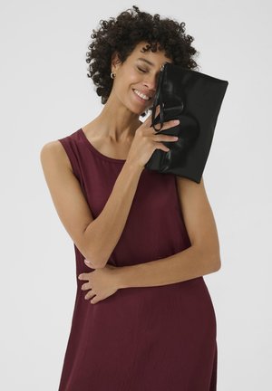 Schwarze Leder-Clutch mit glanzförmiger Oberfläche und Handgelenksriemen, die nah von einer Person gehalten wird, die ein ärmelloses weinrotes Kleid trägt.