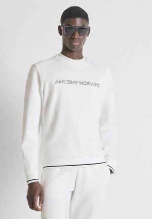 Hombre con sudadera y pantalones blancos de Antony Morato, gafas de sol negras, de pie frente a un fondo liso gris claro.