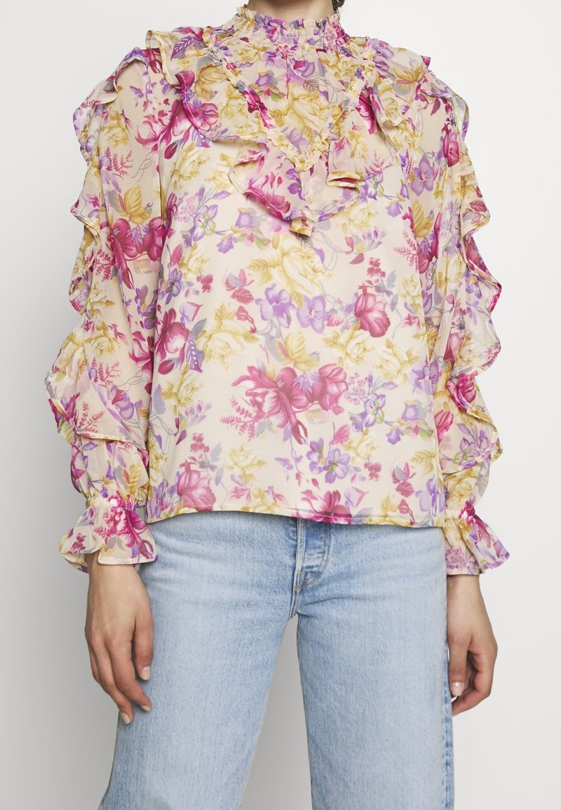 Blouse transparente à motif floral avec des fleurs roses, violettes et jaunes, col montant à volants, manches longues à volants, portée avec un jean bleu clair.