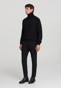 Pullover nero a collo alto con colletto e polsini a coste, abbinato a pantaloni neri slim-fit e scarpe in pelle nere su uno sfondo neutro.
