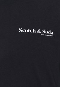 Schwarzes Baumwoll-T-Shirt mit weißem Text, der "Scotch & Soda" und "BORN IN AMSTERDAM" in einer einfachen, fetten Schriftart zeigt.