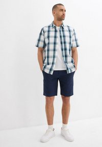 Camicetta a maniche corte a quadri con accenti blu e verdi, base bianca, abbinata a pantaloni corti blu navy e sneakers bianche, in piedi su uno sfondo semplice.