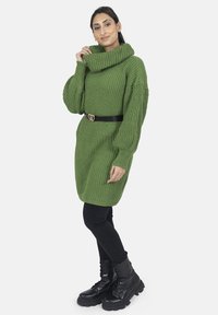 Robe en maille côtelée verte avec col bénitier, ceinturée à la taille avec une ceinture noire, associée à des leggings noirs et des bottes de combat à lacets.