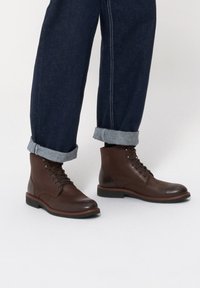 Brązowe skórzane botki za kostkę z okrągłym noskiem, sznurowanym zapięciem i kontrastowym jasnym przeszyciem. Noszone z podwiniętymi ciemnymi dżinsami.