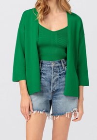 Groene katoenen cardigan met open voorkant, gedragen over een groene ribbel tanktop en hoogwaisted, versleten denim shorts.