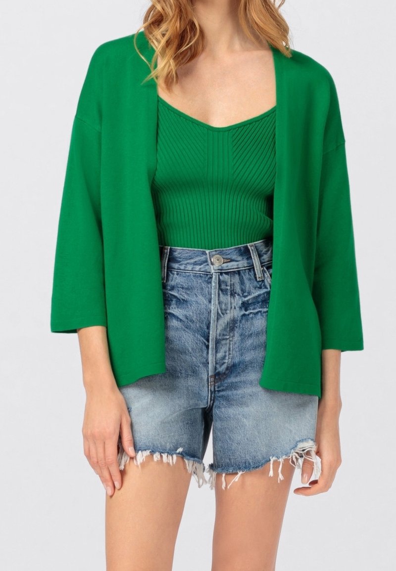 Groene katoenen cardigan met open voorkant, gedragen over een groene ribbel tanktop en hoogwaisted, versleten denim shorts.