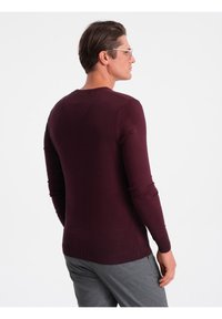 Maglione a manica lunga color bordo, design aderente, scollatura rotonda, polsini e orlo a costine, texture liscia, indossato con pantaloni grigi, visto da dietro.