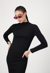 Μαύρο φόρεμα με μακριά μανίκια και υφασμάτινες κάθετες ρίγες, ψηλό λαιμό, εφαρμοστή σιλουέτα, συνδυασμένο με ροζ γυαλιά ηλίου και κρίκους σκουλαρίκια.