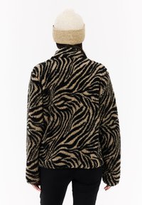 Žena u bež i crnoj jakni sa zebrastrim uzorkom i bež pletenom kapom, okrenuta leđima prema beloj pozadini.