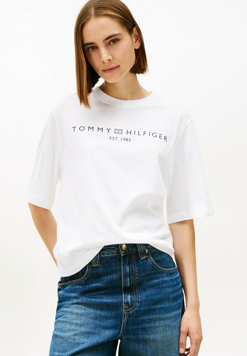 Camiseta blanca de algodón de manga corta con el texto "TOMMY HILFIGER EST. 1985" impreso en azul oscuro, combinada con vaqueros de mezclilla azul.