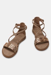 Béžové sandály s křížovými pásky, s širokým kovovým zlatým geometrickým akcentem v přední části a plochou podrážkou s malými nýty.