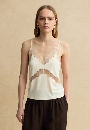 Femme aux cheveux blonds et raides porte un caraco crème orné de dentelle et un pantalon ample foncé sur un fond beige.