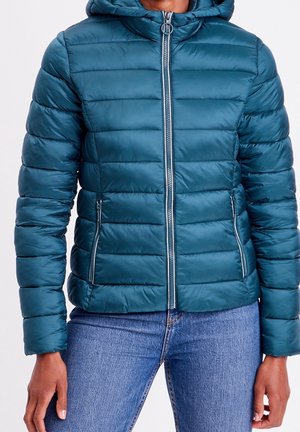 Doudoune matelassée bleu sarcelle avec capuche, fermeture éclair sur le devant et poches zippées portée avec un jean bleu sur une personne debout.