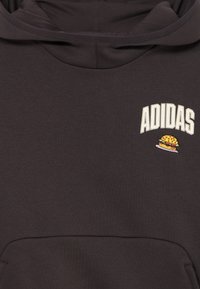 Sudadera gris oscura con bolsillo canguro; presenta el texto "ADIDAS" y un diseño bordado de una hamburguesa en la zona del pecho; textura de tela suave.