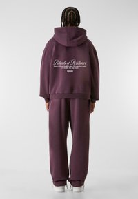 Pflaumenlila übergroßer Hoodie mit einer Vorderasche und weißem Text auf dem Rücken. Kombiniert mit passenden lockeren Sweatpants und weißen Sneakern.