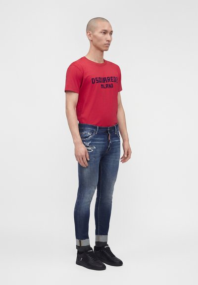 Άνδρας με κόκκινο T-shirt Dsquared2 Milano, φθαρμένο μπλε τζιν με γυρισμένα μανίκια, μαύρα αθλητικά παπούτσια, που στέκεται μπροστά σε λευκό, απλό φόντο.